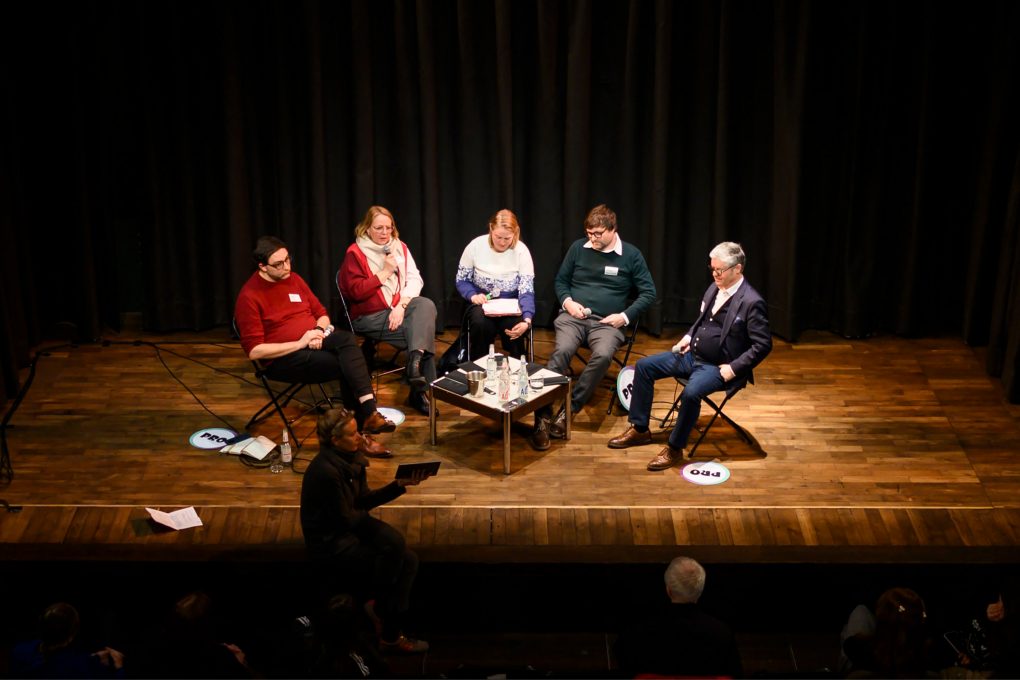 Fünf Personen sitzen auf Stühlen im Halbkreis auf einer Bühne mit Holzboden vor einem schwarzen Vorhang, in der Mitte steht ein kleiner Tisch mit Wasserflaschen und Gläsern
