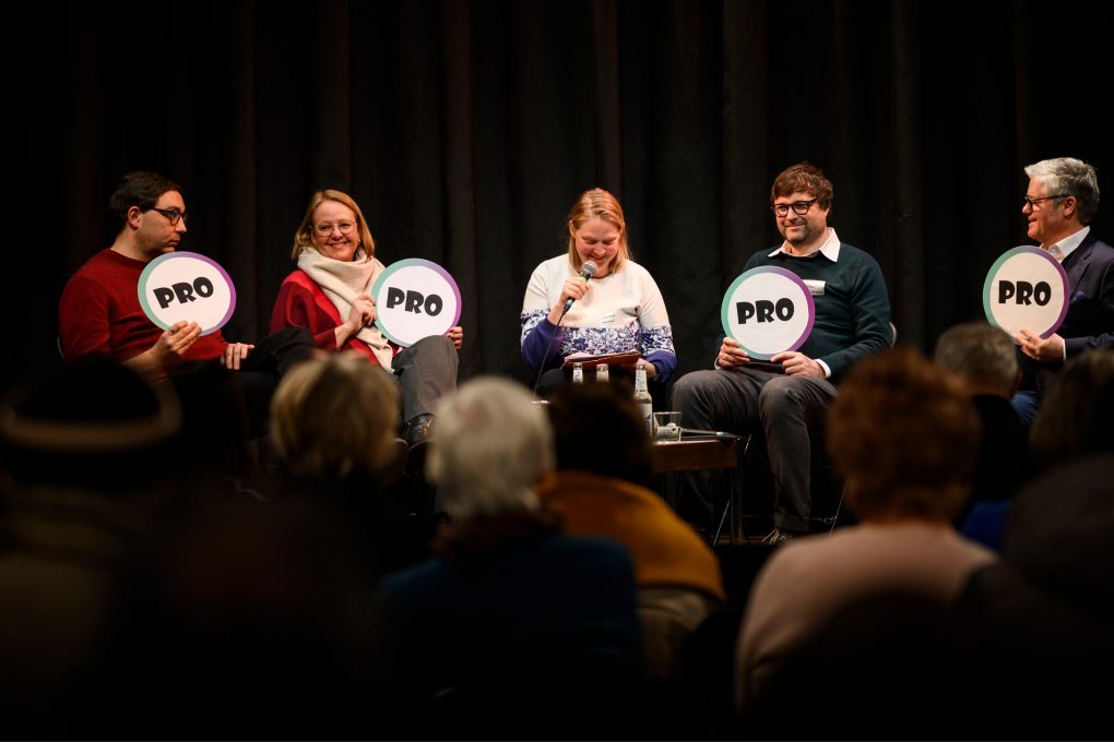 Fünf Personen sitzen auf einer Bühne vor Publikum und halten runde Schilder mit der Aufschrift 'PRO'